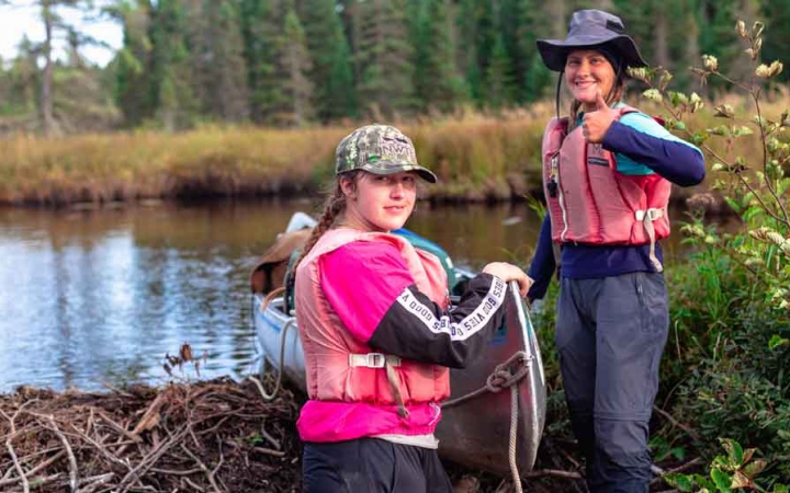 two students smile for a photo while guiding a canoe into water